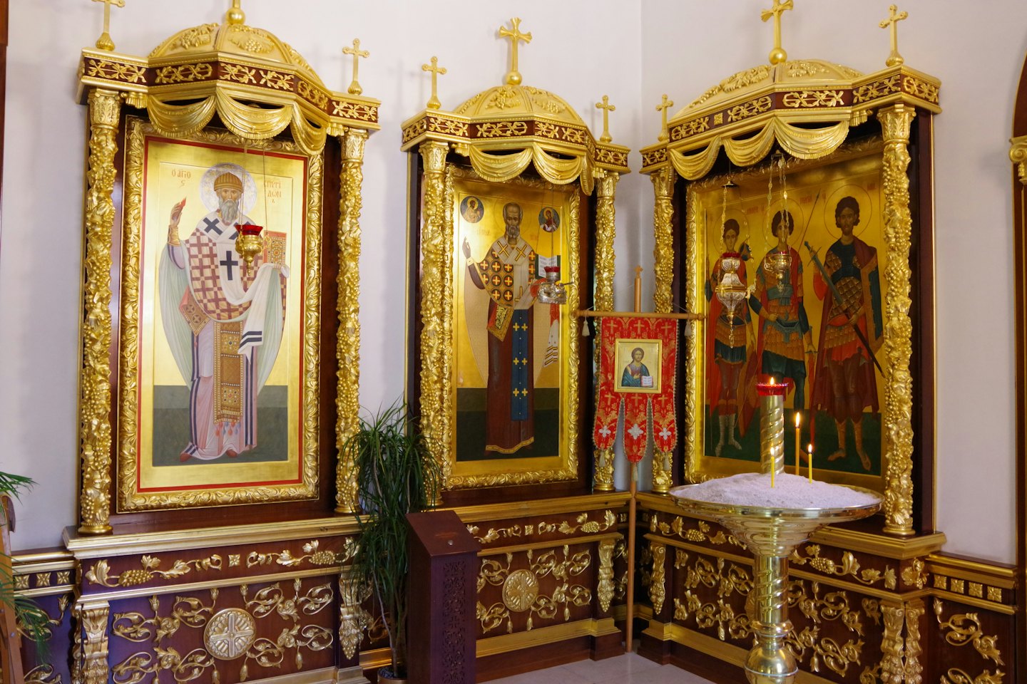 Three golden religious icons displayed near lit candles and plants.